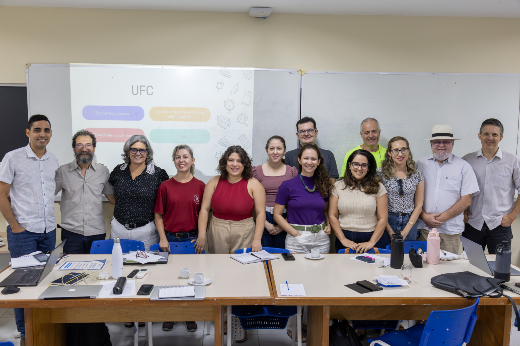 Imagem: Esta imagem mostra um grupo de 14 pessoas, compostas por homens e mulheres adultos, posando para uma foto em uma sala de aula ou reunião. Eles estão de pé, lado a lado, atrás de duas mesas grandes organizadas em forma de U, sobre as quais há cadernos, garrafas, celulares, canetas, papéis e notebooks — sugerindo um ambiente de trabalho ou reunião acadêmica. Ao fundo, há um projetor exibindo um slide com o título "UFC", o que indica que o evento pode estar relacionado à Universidade Federal do Ceará. O fundo é composto por quadros brancos e paredes claras. A maioria das pessoas está sorrindo, aparentando um clima amigável e colaborativo.