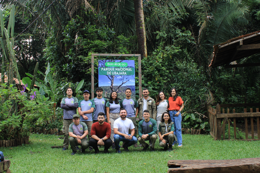 Imagem: Foto de um grupo de 13 pessoas posando ao ar livre em frente a uma placa que diz “Bem-vindos ao Parque Nacional de Ubajara”. O grupo está dividido em duas fileiras: a fileira de trás está em pé e a da frente está agachada. Ao fundo, há vegetação densa, com árvores, plantas tropicais e flores roxas. À direita, vê-se uma estrutura de madeira que parece um pequeno abrigo. O ambiente é verde e natural, transmitindo a sensação de floresta. As pessoas estão sorrindo e vestem roupas casuais, algumas com camisetas do parque.