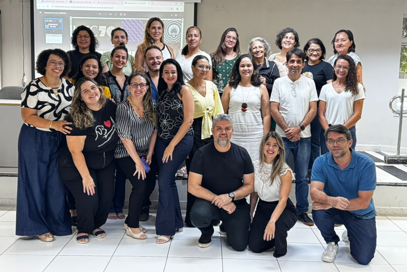 Imagem: A imagem mostra um grupo de aproximadamente 26 pessoas, composto por homens e mulheres, reunidos em um ambiente interno, possivelmente uma sala de reuniões ou auditório. Todos estão posando para a foto, muitos sorrindo, transmitindo um clima de colaboração e acolhimento. Ao fundo, há um telão com projeções que fazem referência ao Processo Estatuinte da Universidade Federal do Ceará (UFC), evidenciado por elementos gráficos e textos parciais visíveis na apresentação. As pessoas estão organizadas em três fileiras: uma em pé, uma agachada ou ajoelhada à frente e outra no meio, ligeiramente abaixada. O grupo representa a comissão setorial do Processo Estatuinte da UFC, formada por docentes, técnico-administrativos e discentes, demonstrando diversidade de gênero, estilos e faixas etárias. O ambiente tem paredes claras, piso branco e uma atmosfera que reforça o caráter institucional, democrático e participativo do momento registrado