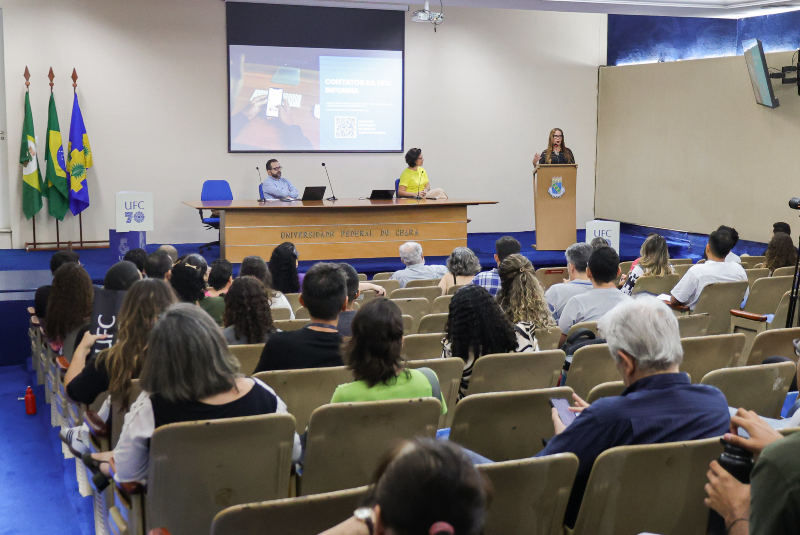 Imagem: A imagem mostra o 1º Encontro da Rede de Comunicadores da UFC (Universidade Federal do Ceará), realizado no auditório da Reitoria da instituição. O ambiente é formal e bem organizado, com diversas pessoas sentadas em poltronas bege, voltadas para o palco onde ocorre a apresentação. A plateia é composta por um público diverso, atento e engajado. No palco, há três pessoas: uma mulher está em pé no púlpito, discursando; outra mulher, vestindo camiseta amarela, está sentada ao centro da mesa principal; e um homem de camisa azul está sentado ao lado esquerdo. Um telão ao fundo exibe uma apresentação com a legenda “Contatos da UFC Informa”, acompanhada de um QR Code. Ao lado do palco, há três bandeiras: do Brasil, do Ceará e da UFC. O espaço é decorado com tons de azul e bege, com destaque para o nome “Universidade Federal do Ceará” gravado na mesa central. A presença de equipamentos como câmera e microfones indica que o evento pode estar sendo registrado ou transmitido. O encontro é uma iniciativa institucional voltada à integração e capacitação dos profissionais de comunicação da universidade.