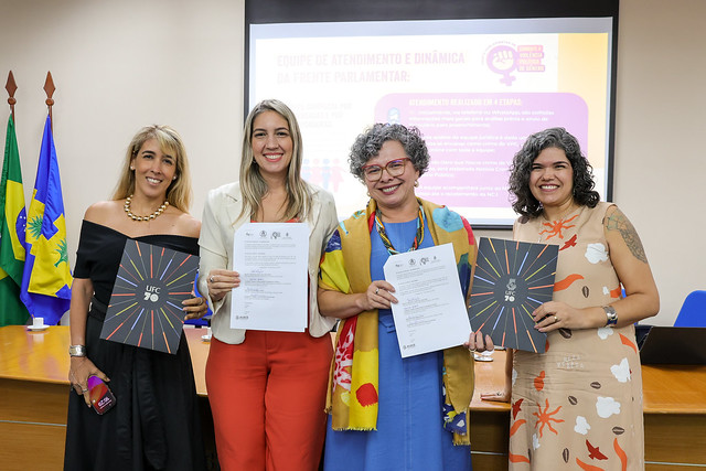Imagem: Quatro mulheres estão lado a lado, sorrindo, em uma sala de reuniões. Elas seguram documentos e pastas da Universidade Federal do Ceará (com a marca UFC 70). Ao fundo, há uma tela de projeção com a apresentação de um slide colorido e três bandeiras: do Brasil, do Ceará e da UFC. As mulheres usam roupas sociais coloridas e estão em pé, próximas a uma mesa com microfones e copos d’água