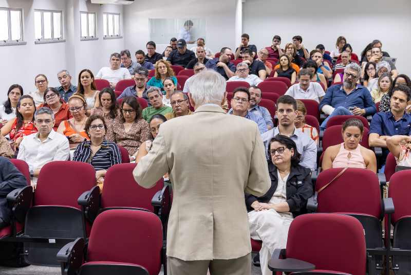 Imagem: A imagem mostra um seminário sendo realizado em uma sala de aula ou auditório. No primeiro plano, está um homem de cabelos grisalhos, vestindo um terno bege, visto de costas — ele parece ser o palestrante ou mediador da atividade. Ele está de pé, falando com o público, que está distribuído em cadeiras vermelhas com apoio para escrita. A plateia está atenta, composta por homens e mulheres de diferentes idades, a maioria com expressões sérias ou concentradas, sugerindo que o tema abordado é de natureza acadêmica ou profissional. As pessoas estão bem-arrumadas, com roupas casuais ou sociais leves. A sala é bem iluminada, com janelas ao longo da parede esquerda e uma cabine de vidro ao fundo, onde duas pessoas podem ser vistas, talvez operando equipamentos de som ou vídeo. O ambiente é formal e parece fazer parte de uma instituição de ensino ou centro de conferências.