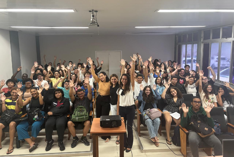 Imagem: A imagem mostra um grupo grande de estudantes reunidos em uma sala de aula ou auditório na unidade didático do Labomar (Instituto de Ciências do Mar da Universidade Federal do Ceará). Os alunos estão sentados em fileiras de cadeiras com braços de apoio, e todos estão voltados para a câmera, sorrindo e com as mãos levantadas, em um gesto de saudação ou entusiasmo coletivo. Na frente da sala, há uma mesa com um projetor, e ao redor dela estão algumas pessoas em pé, que parecem ser professoras, monitoras ou organizadoras da atividade — todas sorrindo, reforçando o clima positivo do encontro. A sala é bem iluminada, com grandes janelas laterais que deixam entrar luz natural, além de lâmpadas fluorescentes no teto. O ambiente sugere um momento de confraternização acadêmica, como o encerramento de um curso, uma recepção para calouros, uma palestra ou outro tipo de evento educativo. A diversidade dos participantes também indica um ambiente inclusivo e acolhedor.