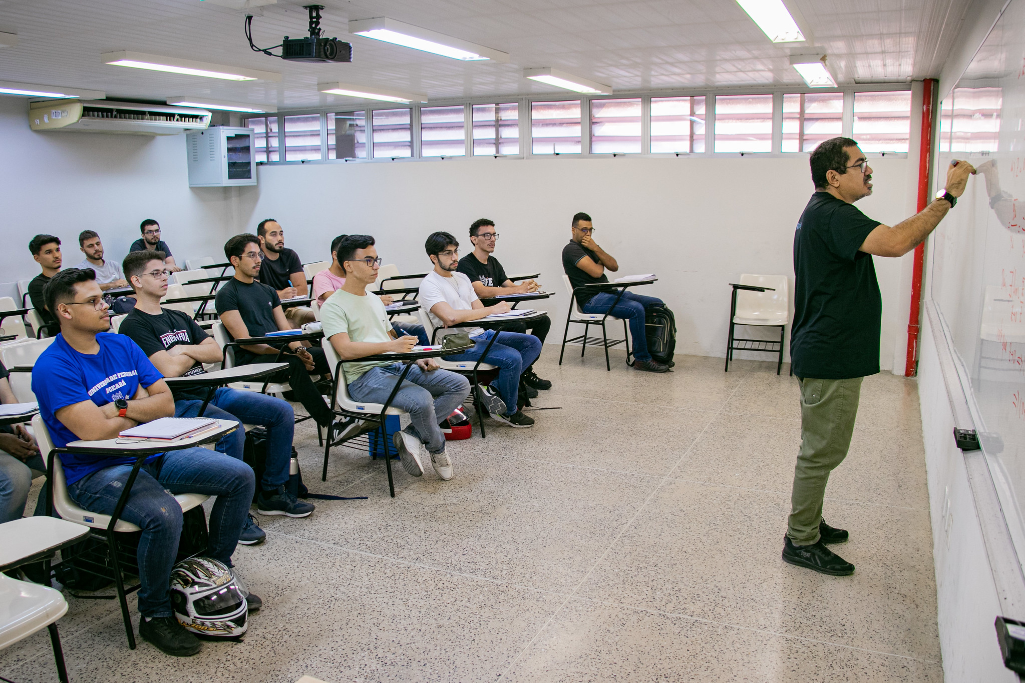Imagem: Sala de aula com cerca de 15 estudantes sentados em carteiras escolares, atentos a um professor que escreve no quadro branco. O ambiente é bem iluminado, com janelas altas e luminárias no teto. Os alunos são majoritariamente jovens e usam roupas casuais. Em primeiro plano, um estudante tem um capacete de moto ao lado da cadeira.