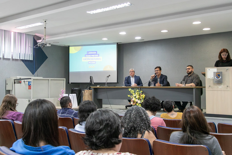 Imagem: foto de pessoas sentadas nas cadeiras do auditório e, à frente delas, um palco com uma mesa onde estão sentados, da esquerda para a direita, Davi Romero, Custódio Almeida e Cícero Miranda