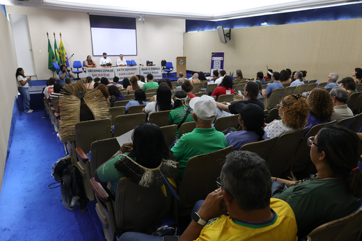 Imagem: auditório durante um seminário, com dezenas de pessoas sentadas acompanhando a programação. À frente, três participantes compõem a mesa de abertura, posicionada em um palco com telão de projeção ao fundo. Em frente à mesa, há faixas estendidas, e, ao lado esquerdo do palco, uma pessoa faz uso da palavra em um púlpito. Ao fundo do auditório, estão as bandeiras do Brasil, do Ceará e da Universidade Federal do Ceará (UFC). A plateia é composta por pessoas de diferentes perfis, algumas utilizando vestimentas e adereços de povos indígenas e movimentos sociais.