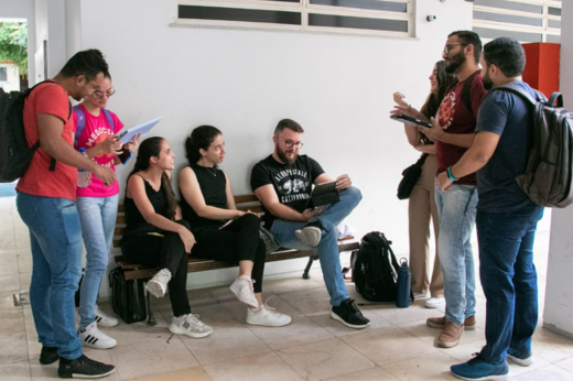 Estudantes interagindo. Três estão sentado no banco, três estão em pé á direita e dois em pé à esquerda