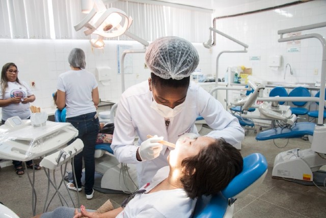 Imagem: Mulher aparece de lado, deitada em cadeira de dentista. Uma dentista, usando jaleco e touca, realiza um procedimento nela. Ao fundo se vêem outras pessoas e equipamentos odontológicos (Foto: Ribamar Neto/UFC)