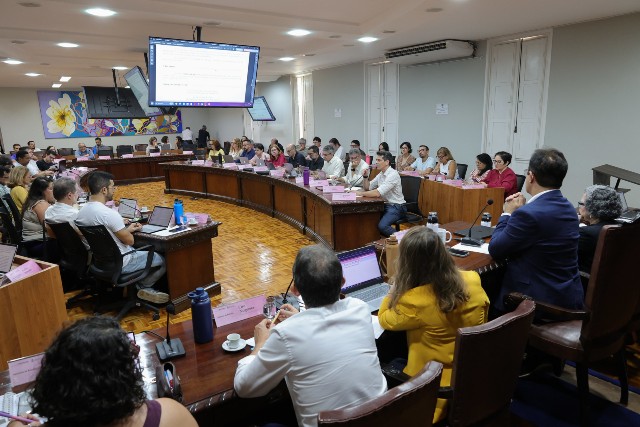 Imagem: Reunião em sala ampla, com dezenas de pessoas sentadas ao redor de mesas em formato circular, usando notebooks, enquanto acompanham apresentações em grandes telas (Foto: Victor Braga/Secom)