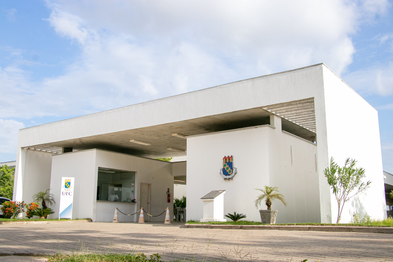 Imagem: A imagem mostra a fachada principal do campus da Universidade Federal do Ceará (UFC) em Russas. A construção tem um design moderno e minimalista, com linhas retas e predominância da cor branca. A estrutura principal é um pórtico retangular que cobre a guarita de entrada, com grandes pilares que sustentam o teto. Na parte frontal, há o brasão da UFC fixado na parede e uma placa informativa logo abaixo. Ao lado, um totem vertical exibe o logotipo da universidade. A entrada possui uma guarita com vidraça para controle de acesso. A área ao redor é bem cuidada, com vasos de plantas ornamentais, arbustos, pequenas palmeiras e flores, conferindo um aspecto acolhedor ao ambiente. O céu está parcialmente nublado, e a iluminação natural valoriza ainda mais a arquitetura limpa e funcional da entrada do campus
