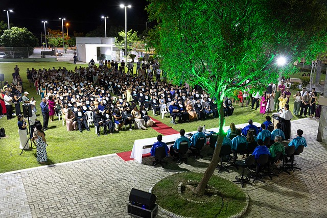 Imagem: Vista aérea de uma cerimônia de formatura ao ar livre durante a noite. Dezenas de pessoas estão sentadas em cadeiras organizadas em fileiras sobre um gramado. À frente, uma mesa coberta com toalha branca e azul fica diante de um pequeno palco onde professores ou autoridades estão sentados.