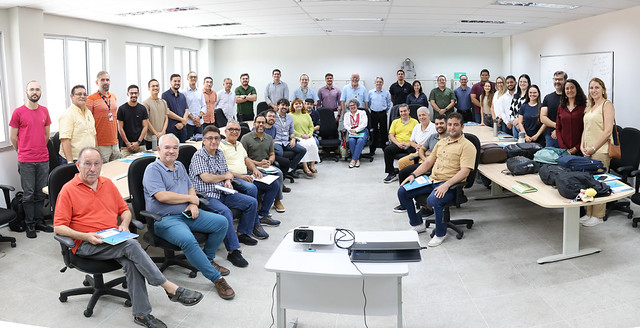 Imagem: Fotografia de um grupo grande de pessoas reunidas em uma sala de reunião ampla e bem iluminada, com paredes brancas e janelas laterais. Cerca de 30 a 40 participantes, entre homens e mulheres de diferentes idades, estão distribuídos entre cadeiras e em pé ao fundo, muitos sorrindo e olhando para a câmera. Na parte frontal da imagem, há uma mesa com um projetor e um notebook. As pessoas vestem roupas casuais e de trabalho, como camisas polo, camisetas e calças. O ambiente sugere um encontro institucional ou reunião de equipe.