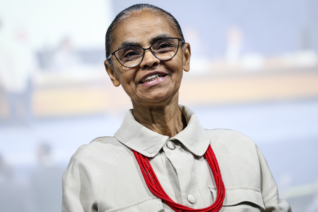 Imagem: Marina Silva em um retrato de meio corpo, posicionada de frente para a câmera e com uma leve inclinação da cabeça. Ela sorri suavemente, transmitindo uma expressão acolhedora e serena. A pessoa usa óculos de armação fina e tem os cabelos grisalhos presos para trás de forma discreta. Está vestida com uma camisa de tom bege claro, de mangas compridas, com gola estruturada e botões frontais. Como acessório de destaque, usa um colar longo composto por várias voltas de contas vermelhas, que contrastam com a neutralidade da roupa.