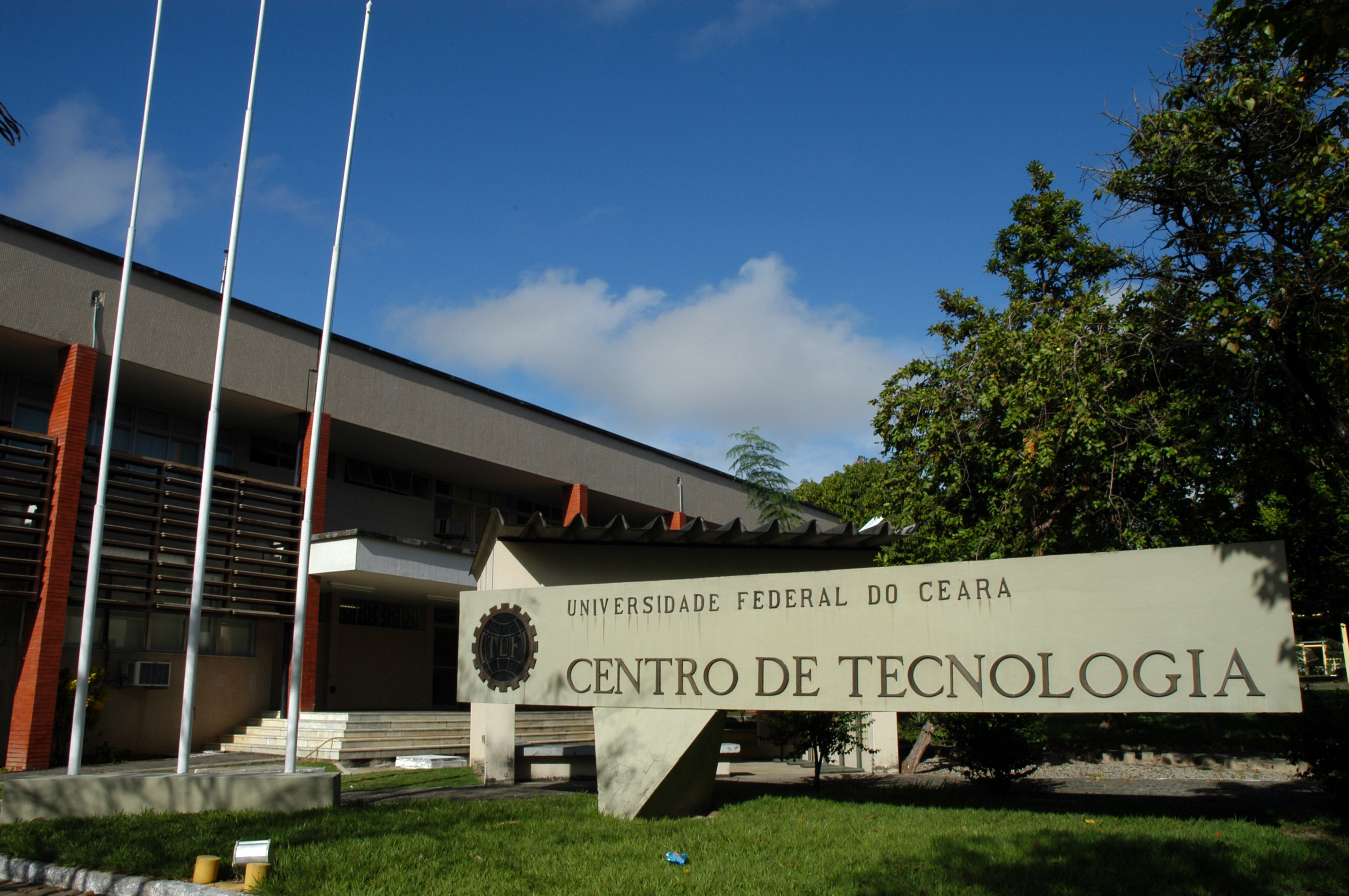Imagem: Placa sinalizadora do Centro de Tecnologia da UFC, localizada na frente do prédio principal e fincada em gramado verde, com árvores do lado direito e três mastros do lado esquerdo, com visão para a escada de acesso.