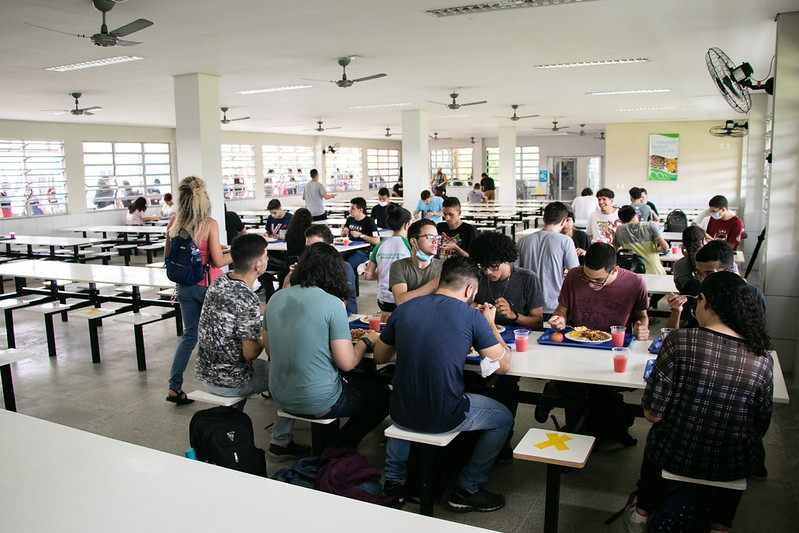 Imagem: Foto interna do Restaurante Universitário do Campus do Pici da UFC, com mesa à direita ocupada por nove estudantes, uma aluna de blusa rosa e mochila preta caminhando no corredor e carregando uma bandeja, e mais três mesas ao fundo do refeitório, à direita, com alunos almoçando.
