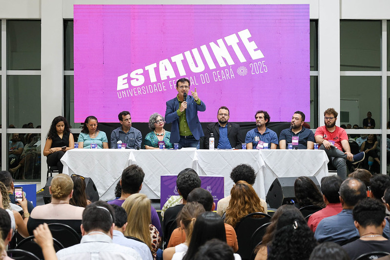 Mesa de abertura do I Congresso da Estatuinte da UFC com nove pessoas sentadas e, ao centro, em pé, o reitor Custódio Almeida discursa para plateia presente. Atrás do reitor, há um painel de cor roxa com a frase, na cor branca “Estatuinte Universidade Federal do Ceará 2025”.