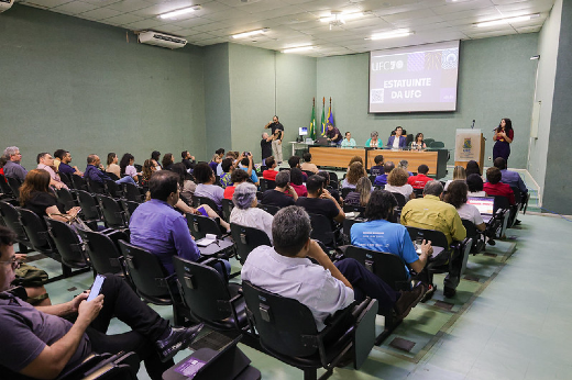 Imagem: foto de um auditório com pessoas sentadas am cadeiras e, na frente, um palco com uma mesa e algumas autoridades da UFC