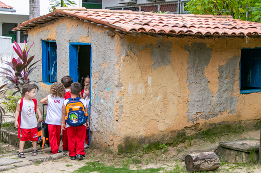 Imagem: Foto das crianças do NDC CAp, vestidas com farda branca e vermelha, brincando de costas em uma casinha de taipa que faz parte do parque infantil. (Foto: Álvaro Graça Jr./UFC Informa)