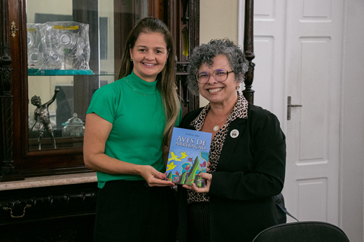 Imagem: A secretária Yrwana Albuquerque e a vice-reitora Diana Azevedo seguram juntas um exemplar do livro Aves de Arribação, de Antônio Sales.