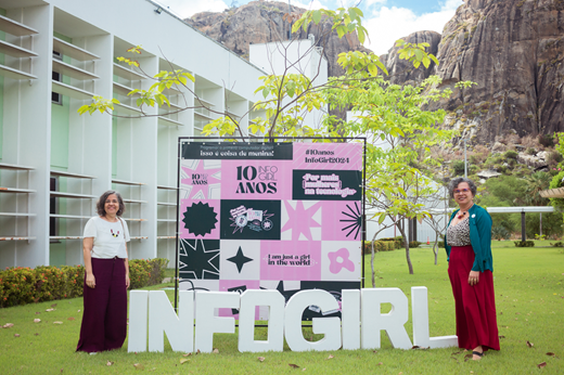 Imagem: As professoras Áurea Holanda e Diana Azevedo posam nos jardins do Campus de Quixadá. Entre elas, está um banner e o letreiro do evento INFOGIRL. 