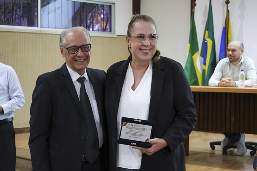 Prof. Tomaz Nunes e Prof Cláudia do Ó Pessoa posam durante a solenidade; ela segura placa de homenagem