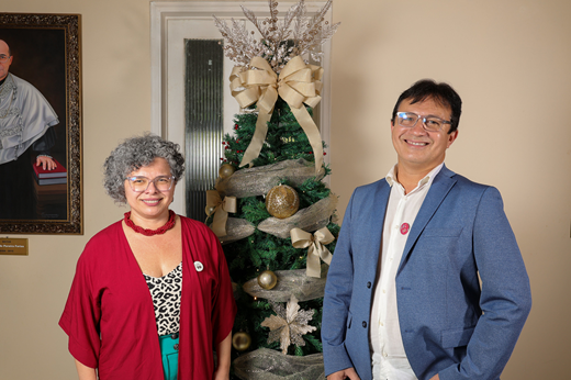 Imagem: A vice-reitora Diana Azevedo e o reitor Custódio Almeida posam em frente a árvore de Natal instalada na Galeria de Ex-Reitores; ela traja roupa vermelha e ele, terno azul.