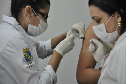 Imagem: Foto de mulher vestida de jaleco e com mascara cirúrgica aplicando vacina no braço de uma pessoa