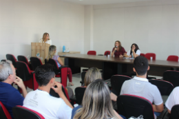 Imagem: Foto do auditório a partir do fundo, onde se vê servidores sentados de costas e, na mesa do evento, Profª Maria Elias e Profª Marilene Feitosa
