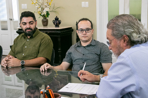 Imagem: Foto do reitor Cândido Albuquerque assinando um documento ao lado dos professores Sandro Vagner e Wellington Franco sentados na mesa