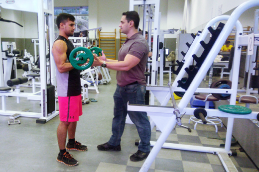 Imagem: Jovem fazendo musculação na academia; erguendo uma barra com auxílio de um instrutor (Foto: Divulgação/IEFES)