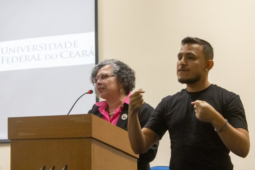 Imagem: Foto com close da vice-reitora Diana Azevedo fazendo um discurso no púlpito, ao lado de um intérprete de Libras