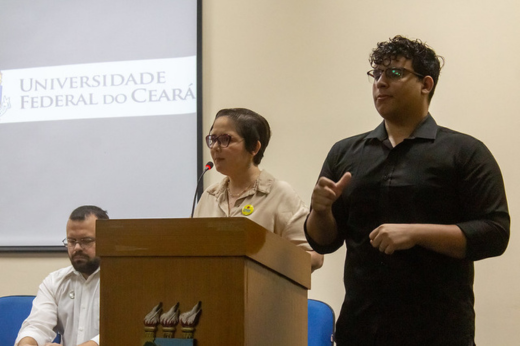 Imagem: Foto da servidora Elaine Teixeira discursando no púlpito, ao lado do intérprete de Libras