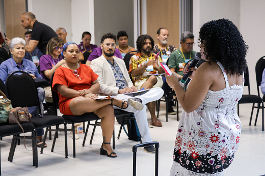 A imagem mostra uma pessoa com cabelo cacheado, vestindo um vestido branco com estampa floral, falando ao microfone em frente a um grupo de pessoas sentadas em cadeiras. A oradora está de costas para a câmera. Os participantes estão atentos, com expressões diversas. À esquerda, uma mulher de vestido vermelho e cabelos presos observa com atenção, enquanto outras pessoas, de diferentes idades, gêneros e etnias, estão sentadas em fileiras. No fundo, há uma porta aberta e mais pessoas interagindo. O ambiente é iluminado, e parece ser uma sala de reuniões ou auditório (Foto:  Ribamar Neto/UFC)