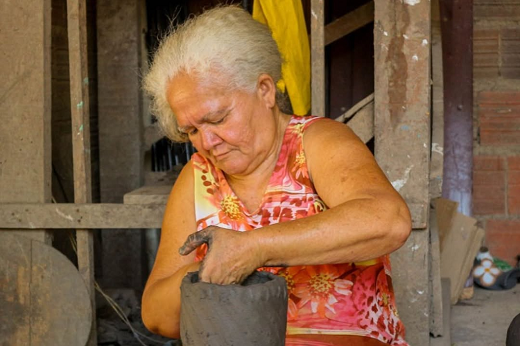 Imagem: Na imagem, vemos uma mulher idosa moldando uma peça de barro em um ambiente rústico. Ela tem cabelos curtos e brancos e veste uma blusa estampada com tons de rosa e laranja. Suas mãos estão concentradas no trabalho artesanal, enquanto amassa e molda a argila. Ao fundo, há uma estrutura de madeira e alguns objetos desfocados, sugerindo um ateliê ou espaço de trabalho típico de artesãos. O cenário transmite dedicação e conexão com a tradição do artesanato (Foto: Centro de Design do Ceará) 