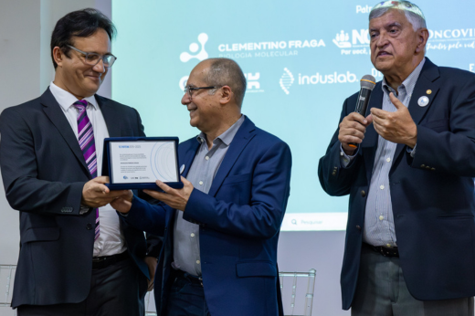 Prof. Odorico falando ao microfone. Custódio (reitor) e Jesualdo (ex-reitor) se olhando enquanto seguram a placa dos homenageados na solenidade de 10 anos do NPDM