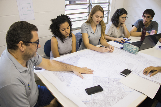 Imagem: Um professor e um grupo de alunos e alunas sentados em volta de uma mesa, analisando um projeto em papel sobre a superfície plana, em uma sala de laboratório (Foto: Ribamar Neto/UFC)