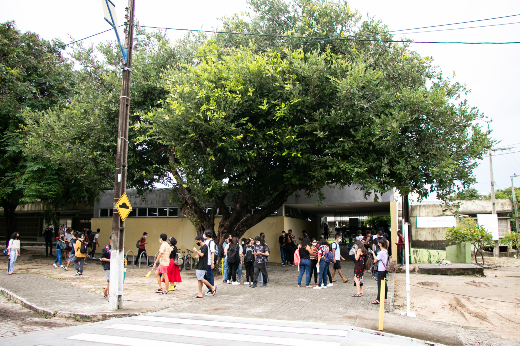 Imagem: A imagem mostra um grupo de estudantes reunidos em frente ao restaurante Universitário do Campus do Pici.Há uma grande árvore no centro da cena, e, em frente dela, há um poste de energia com fios. Há também uma faixa de pedestres no primeiro plano.