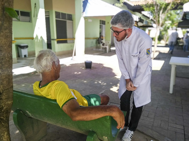 Imagem: um senhor de camisa amarela está sentado em um banco de costas para a foto. Em pé, um jovem de jaleco brando e touca conversa com ele (Foto: Luana Oliveira/UFC)