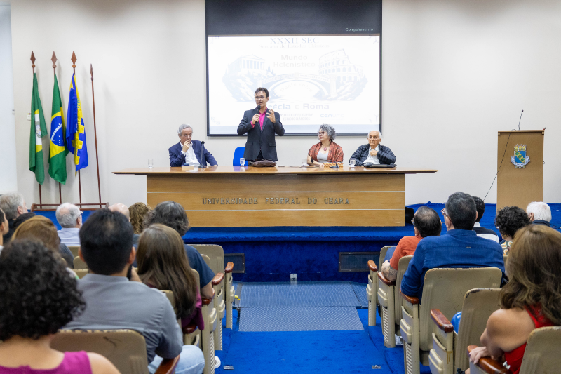 Imagem: Foto do reitor Custódio Almeida em pé, os convidados sentados e o público do auditório da Reitoria no primeiro plano