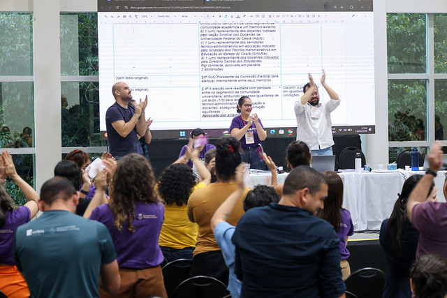 Em um auditório, várias pessoas estão na plateia aplaudindo de pé. No palco, um homem adulto careca de blusa azul marinho, uma mulher branca de óculos de grau e blusa roxa e um homem de camisa branca tambḿe estão aplaudindo de pé. Eles estão de frente para a plateia. Atrás deles, um telão expõe um documento em formato de tabela com três colunas.