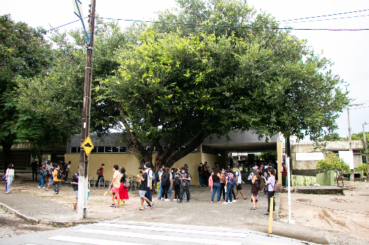 Imagem: pessoas circulam na entrada de um dos prédios do campus do Pici, com árvores frondosas próximas e um poste de energia elétrica à frente