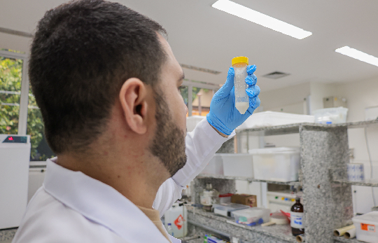 Imagem:Foto de pesquisador segurando um frasco transparente com o repelente. O pesquisador é homem, branco. Usa uma bata de laboratório e está de lado, quase de costas. Ele usa uma luva azul para segurar o frasco. Ao fundo, se vê bancada de laboratório (Foto: Ribamar Neto/UFC Informa)