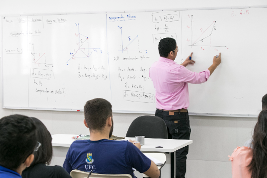 Imagem: estudantes sentados em carteiras escolares, de costas, olhando para um professor (também de costas). O professor está diante de um quadro, no qual estão escritas várias fórmulas e equações matemáticas (Foto: Viktor Braga/UFC Informa)  