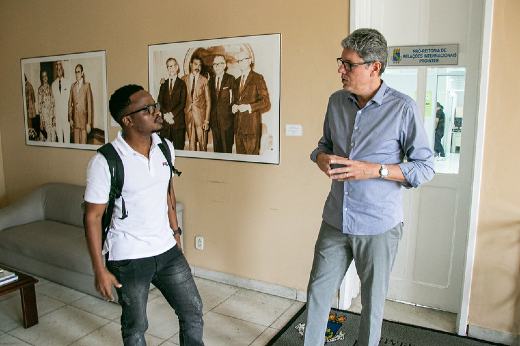 Imagem: Dois homens, um negro com blusa branca e outro branco e alto com blusa cinza, conversam na frente de uma porta, em ambiente fechado com fotografias na parede