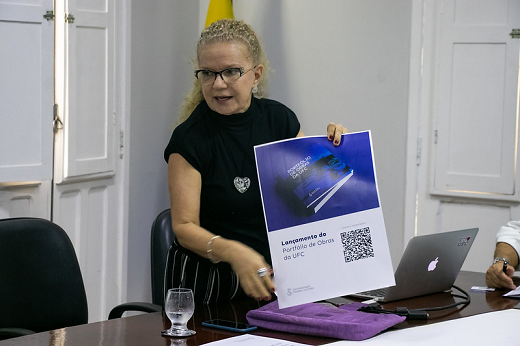 Imagem: Profª Cláudia Buhamra segura cartaz de lançamento do livro, com QR Code para acesso ao e-book