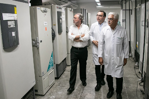 Imagem: homens de jaleco branco caminham em ambiente de laboratório ao redor de grandes máquinas de freezers brancas