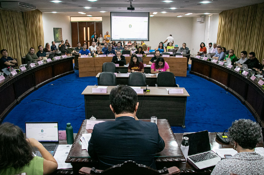 Imagem: A imagem mostra uma reunião em uma grande sala, com várias pessoas sentadas ao redor de uma mesa oval ou em formato de U. Há uma tela de projeção ao fundo com o logotipo de uma instituição, e as pessoas parecem estar participando de uma sessão ou audiência formal, com algumas usando laptops para tomar notas ou acompanhar o evento. Nas mesas, há placas de identificação para cada participante. O ambiente parece organizado, e o evento parece ser uma reunião ou conferência de caráter oficial, possivelmente em um contexto acadêmico ou governamental.