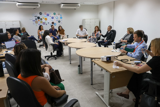 Imagem: A imagem mostra um grupo de pessoas sentadas em uma sala, participando de uma reunião. O ambiente é iluminado e moderno.