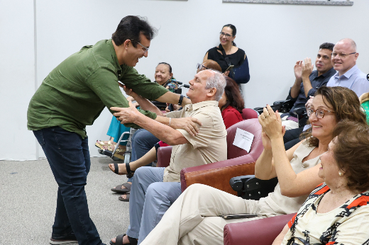 Imagem: homem de blusa social verde, cabelos lisos curtos e óculos curva-se para a frente para cumprimentar um senhor de idade, careca e com deficiência nos olhos, sentado em uma das poltronas de um auditório