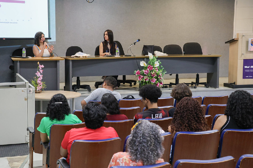 Imagem: A imagem mostra uma apresentação em um auditório. No palco, duas mulheres estão sentadas à mesa em frente a um grupo de pessoas. Uma delas fala com um microfone, enquanto a outra está atenta. Há arranjos de flores no palco, e o público, composto por várias pessoas, assiste atentamente. Um slide está parcialmente visível no projetor à esquerda.